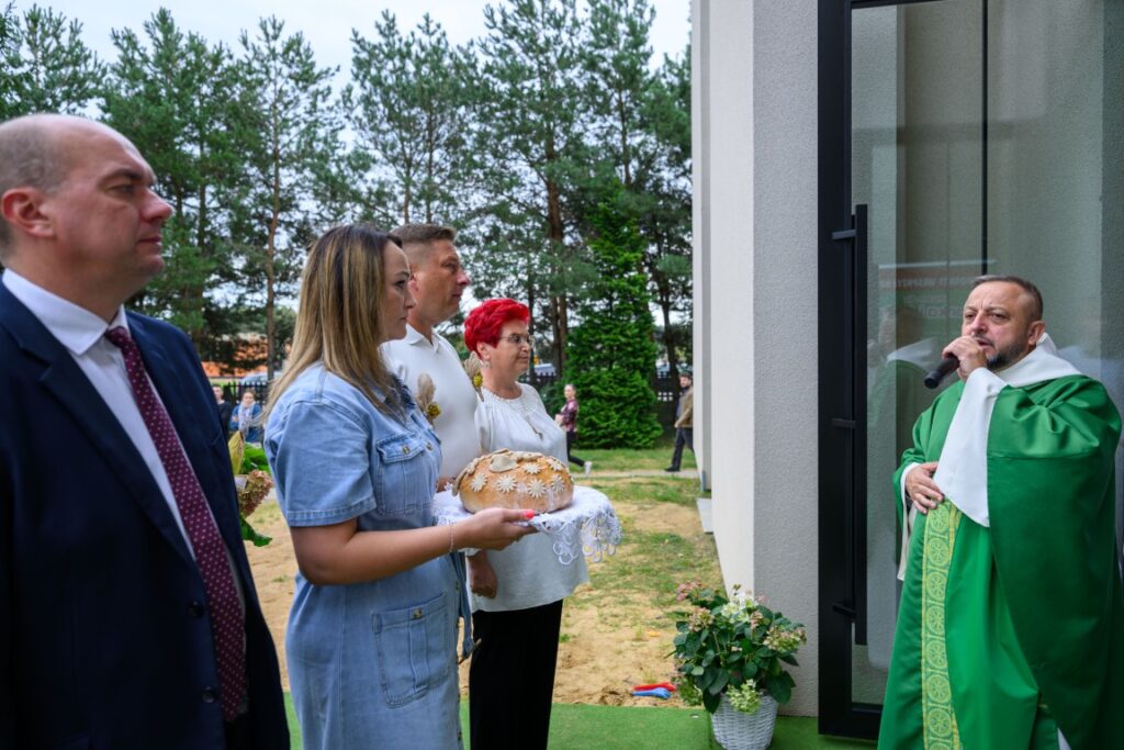 Grupa ludzi stoi na zewnątrz budynku przy księdzu w zielonej szacie. Kobieta trzyma chleb na białej serwetce. W tle widać drzewa i trawnik.