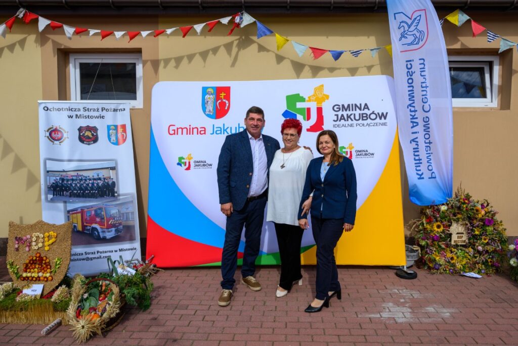 Trzy osoby stoją przed kolorowym banerem Gminy Jakubów. Tło ozdobione jest girlandą i plakatami straży pożarnej. Na ziemi widać dekoracje z kwiatów.