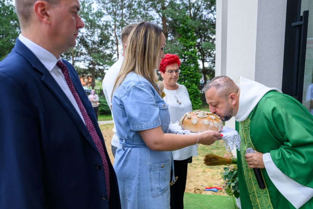 Grupa osób podczas ceremonii; ksiądz w zielonym ornacie całuje chleb trzymany przez kobietę w niebieskiej sukience. Tło zielone drzewa.