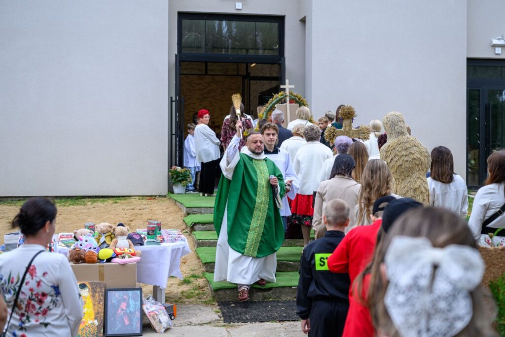 Kapłan w zielonej szacie prowadzi procesję do kościoła. Tło stanowi szare, proste elewacje budynku. Obok stoły z zabawkami i obrazami.
