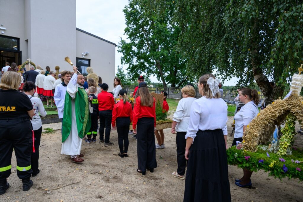 Grupa ludzi w odświętnych strojach przed budynkiem. Zielony trawnik i białe ściany w tle. Widoczne są także dekoracyjne wieńce.