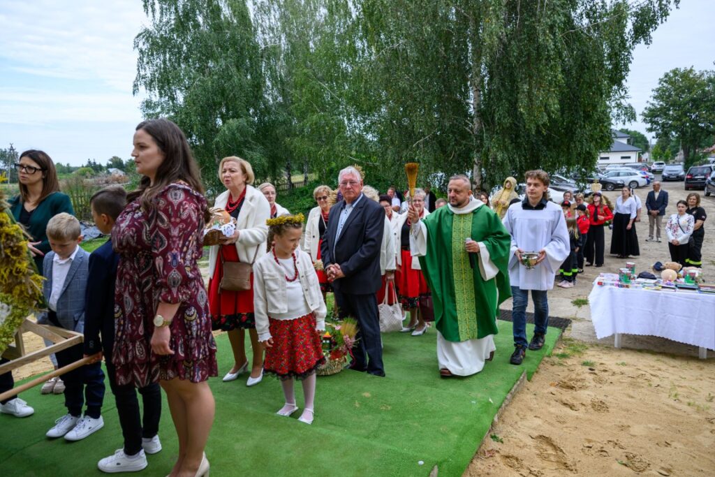 Grupa ludzi w strojach ludowych idzie po zielonym dywanie. W tle drzewa i stolik z białym obrusem. Osoba w zielonym ornacie prowadzi procesję.