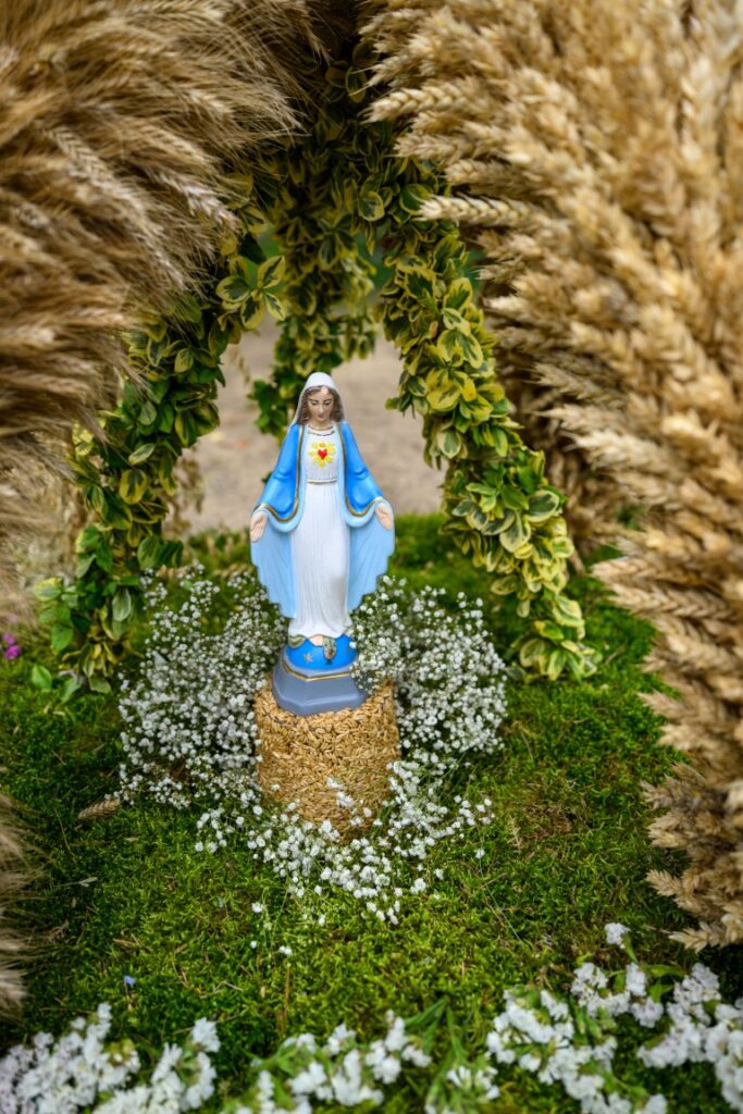 Figurka Matki Boskiej w niebieskiej szacie, otoczona zielonymi liśćmi i białymi kwiatami, na tle traw i kłosów.