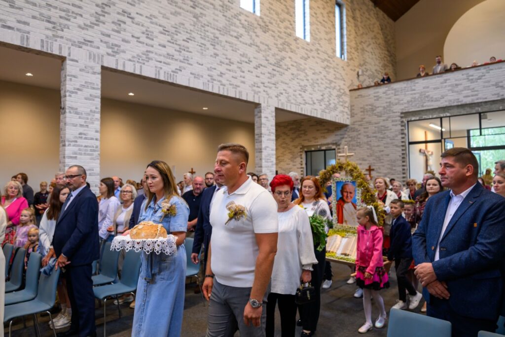 Ludzie w kościele niosą chleb na białej serwecie. Ściany są jasnoszare, a wewnątrz jest tłum w eleganckich ubraniach. W tle widoczna jest dekoracja z krzyżem.