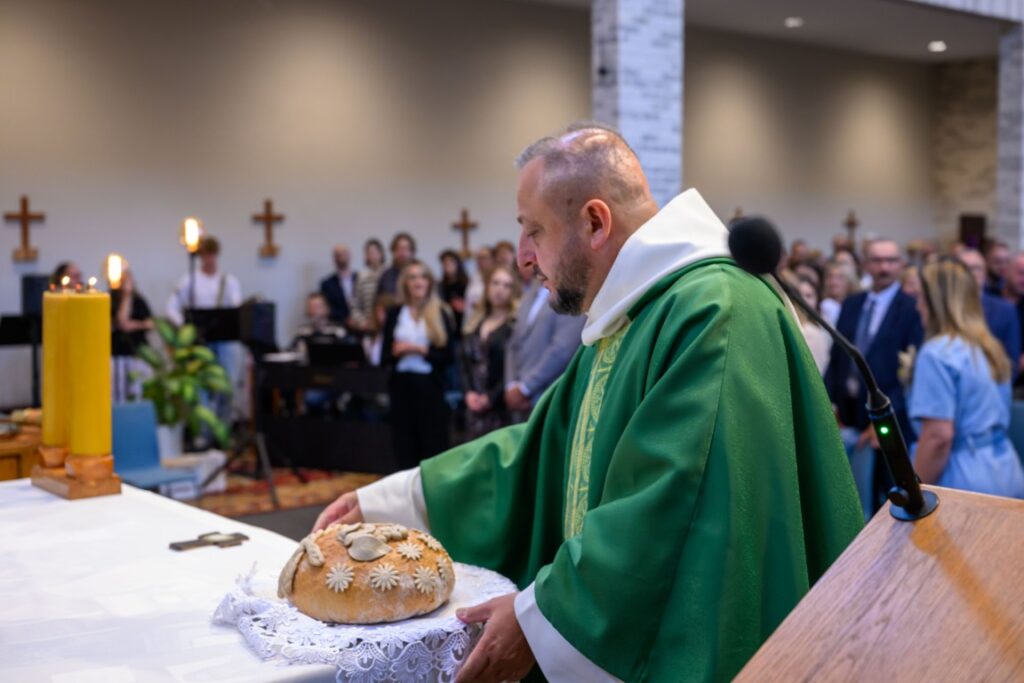 Duchowny w zielonym ornacie trzyma chleb na ołtarzu. W tle widać ludzi i krzyże na ścianie. Kolory są stonowane.