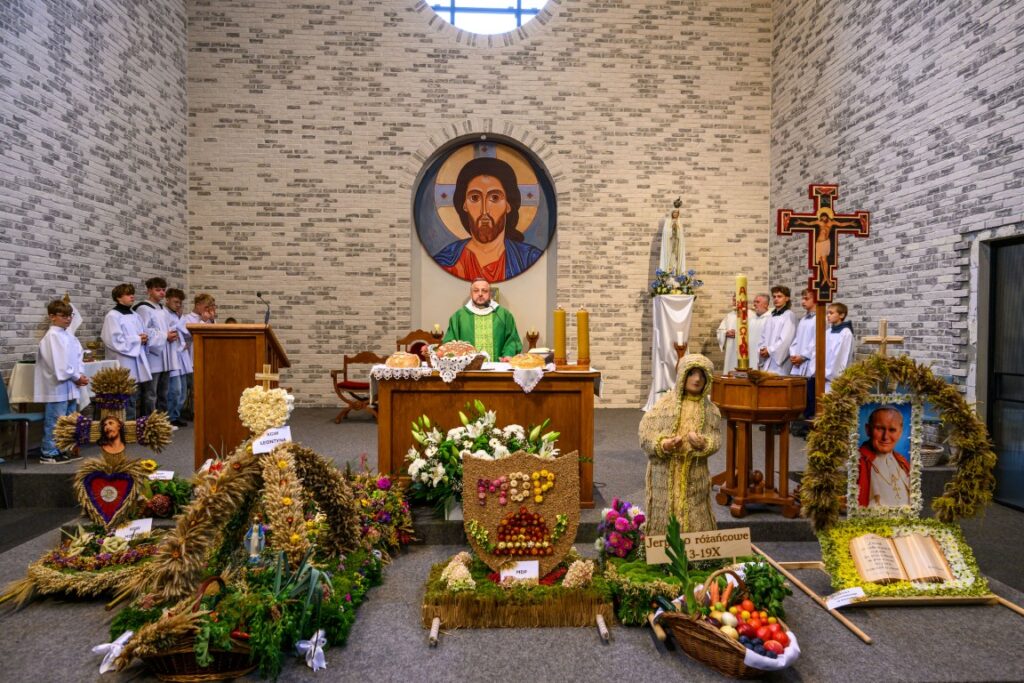 Ołtarz kościelny z jasełką, księdzem w zieleni, ołtarz ozdobiony kwiatami i koszami plonów. Tło z jasnej cegły.