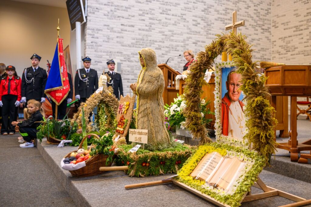 Na zdjęciu widać dożynkowe dekoracje, z wieńcami i plonami, w kościele. Kolory są naturalne: zielenie, złoto, brązy. W tle znajduje się grupa osób w mundurach i ceglana ściana.