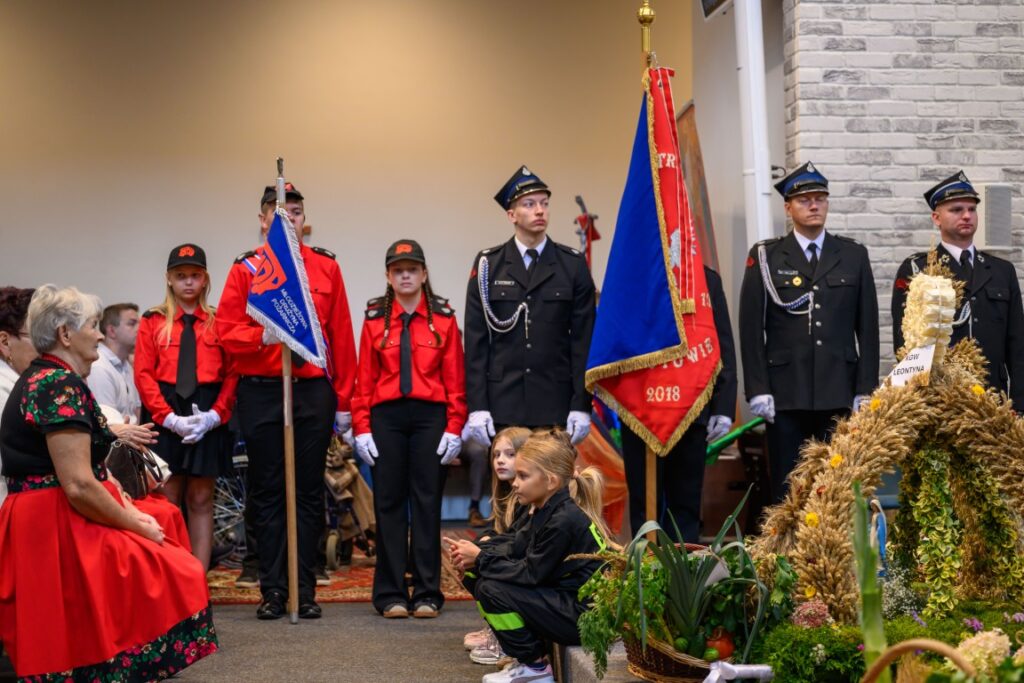 Ceremonia z udziałem strażaków w czerwonych i czarnych mundurach, trzymających flagi. W tle ściana z szarych cegieł. Na pierwszym planie kompozycja z dożynkami.
