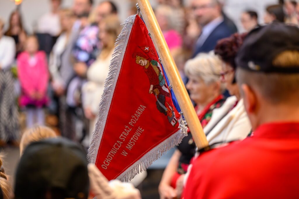 Zdjęcie przedstawia czerwony sztandar Ochotniczej Straży Pożarnej z napisem, trzymany przez mężczyznę w czerwonym stroju, na tle tłumu. W tle nieostry tłum ludzi. Kolory dominujące to czerwony i szary.