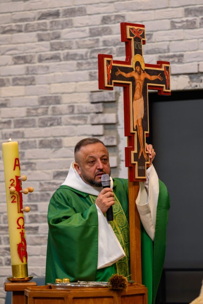 Na zdjęciu znajduje się mężczyzna w zielonej szacie liturgicznej. Trzyma krzyż z czerwonymi elementami i mikrofon. W tle szara ceglasta ściana.
