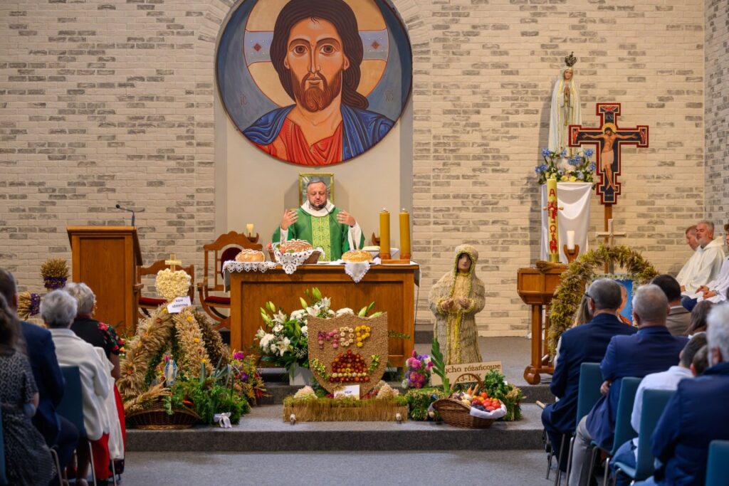 Na tle białej ceglanej ściany w kościele kapłan w zielonym ornacie celebruje Mszę. W tle duża ikona Jezusa, statuetka i kwiaty. Wokół ołtarza ozdoby z plonów i kwiatów.
