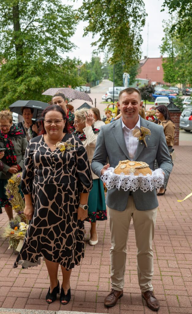 Dwóch ludzi w eleganckich strojach przed budynkiem; mężczyzna trzyma chleb. W tle grupa osób z parasolami na tle zieleni.