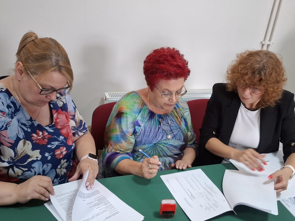 Trzy kobiety przy stole przeglądają dokumenty. Mają kolorowe ubrania, a tło jest białe. Na stole leży czerwony stempel.