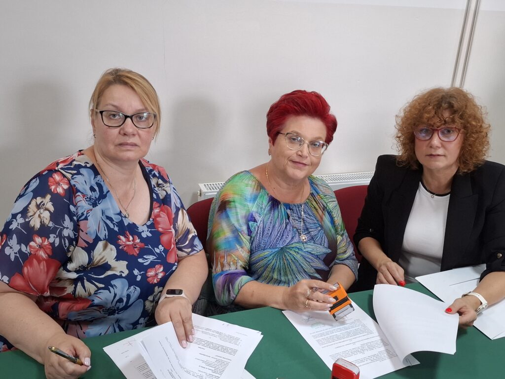 Trzy kobiety siedzą przy stole z zielonym obrusem, przeglądając dokumenty. Ubrane są w kolorowe stroje, a tło jest jasnoszare. Jedna z nich trzyma pieczątkę.