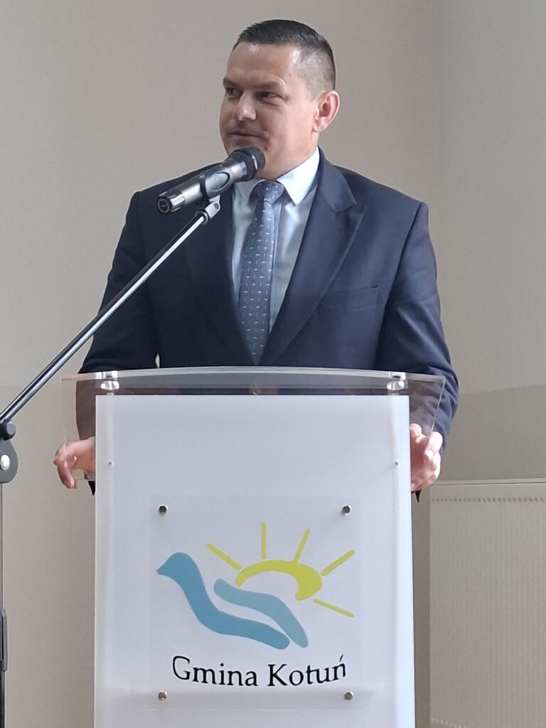 Mężczyzna w garniturze mówi do mikrofonu stojąc za mównicą z logo Gminy Kotuń. Tło jest jasne i neutralne.