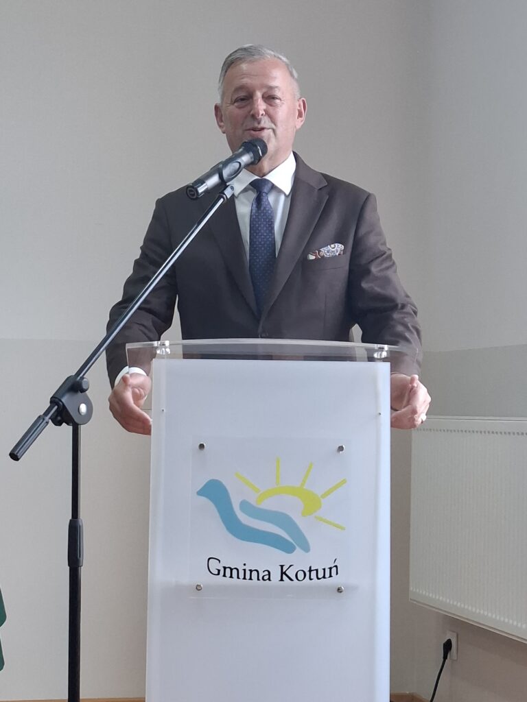 Mężczyzna w garniturze przemawia do mikrofonu stojąc za przezroczystą mównicą z logo Gminy Kotuń. Tło jest jasne, z beżową ścianą i kaloryferem. Strój mężczyzny jest ciemny z kolorową poszetką.