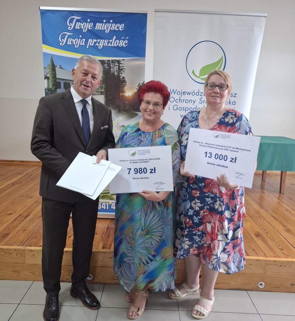 Trzy osoby na podium trzymają tablice z kwotami. Kobiety w kolorowych sukienkach, mężczyzna w garniturze. Tło z banerami promocyjnymi.