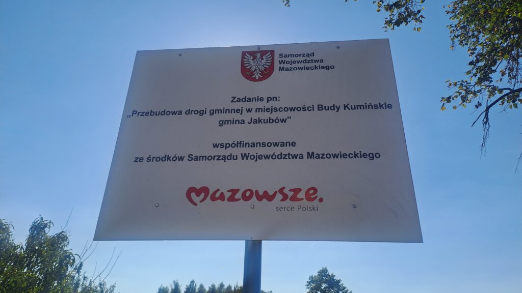 Tablica informacyjna na tle nieba, dotycząca przebudowy drogi w Budach Kumińskich. Widoczne logo Mazowsza. Zielone gałęzie po bokach.