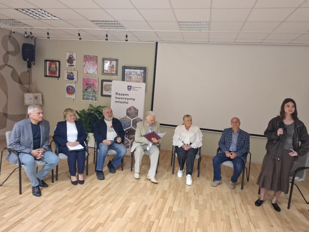 Grupa ludzi siedzi na krzesłach w jasnym pomieszczeniu z drewnianą podłogą. Na ścianie wiszą kolorowe obrazy, obok biały baner z napisem Razem tworzymy miasto. Kobieta z mikrofonem stoi z prawej strony.