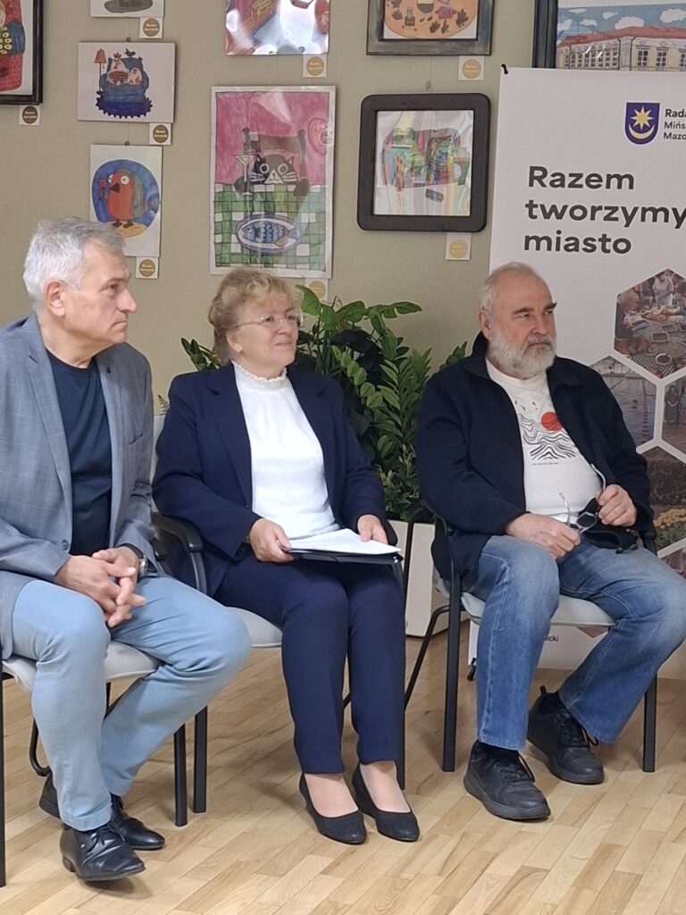Trzy osoby siedzą na krzesłach w jasnym pokoju z drewnianą podłogą. W tle widać kolorowe obrazy oraz rośliny. Po prawej stronie stoi biała tablica z napisem.