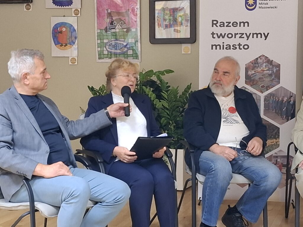 Na zdjęciu są trzy osoby siedzące w jasnym pomieszczeniu, jedna trzyma mikrofon. W tle wiszą kolorowe obrazy i rośliny. Obok widoczny jest roll-up z napisem Razem tworzymy miasto.