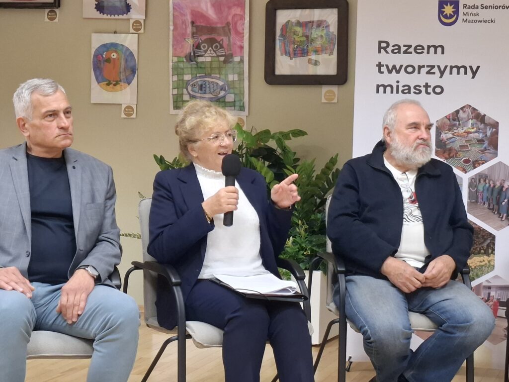 Trzy osoby siedzą na krzesłach, kobieta trzyma mikrofon. Tło z roślinami i plakatem Razem tworzymy miasto. Ściana ozdobiona obrazkami.