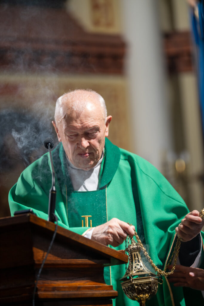 Kapłan w zielonym ornacie trzyma kadzielnicę, woń dymu unosi się wokół niego. W tle rozmyte wnętrze kościoła. Na przodzie drewniana ambona z mikrofonem.