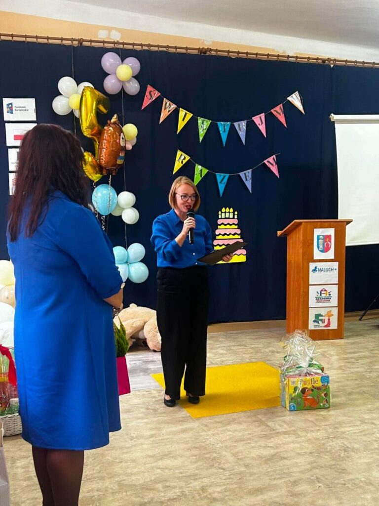 Osoba w niebieskiej bluzce mówi do mikrofonu na tle kolorowych balonów i girland. Obok widoczny jest prezent oraz podium. Dekoracje nawiązują do święta lub uroczystości.