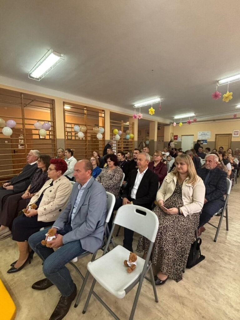 Grupa ludzi siedzi na krzesłach w jasnym pomieszczeniu. Ściany ozdobione są balonami i papierowymi kwiatami. W tle drewniane drabinki gimnastyczne.