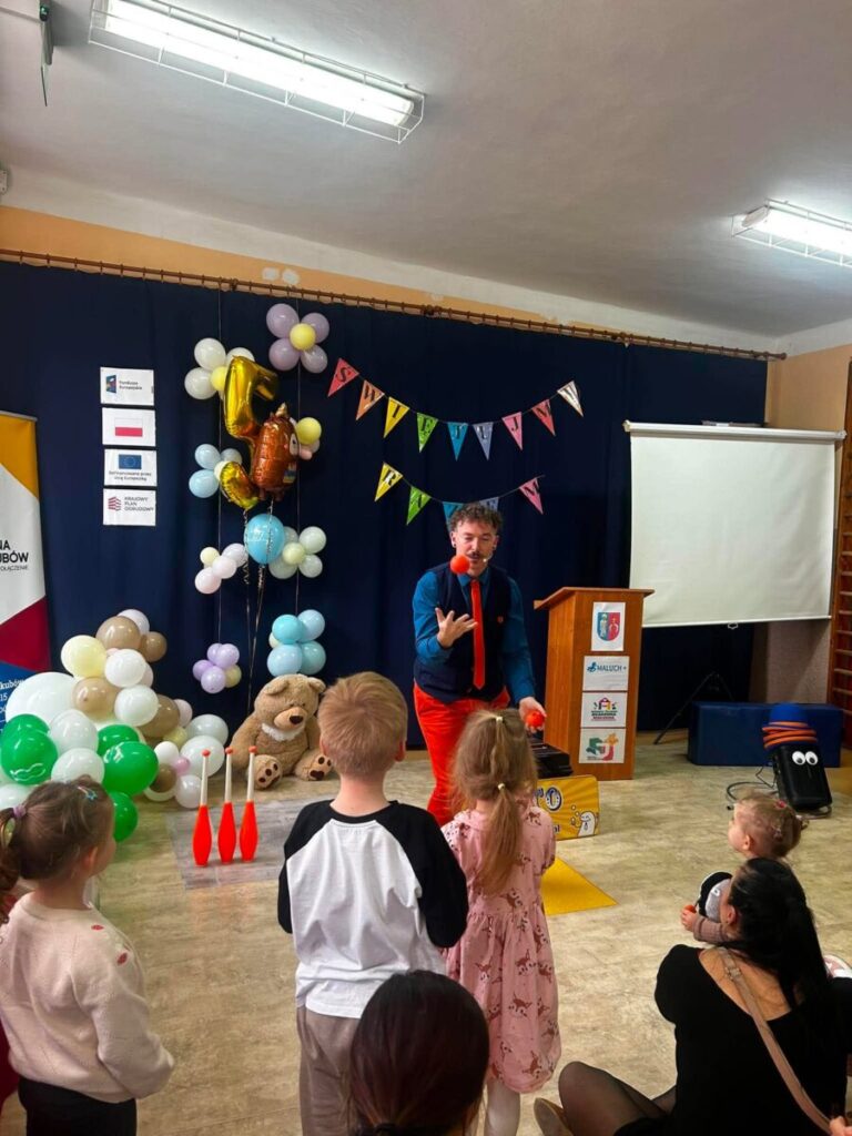 Przedstawienie klauna z balonami i dziećmi na tle granatowej kurtyny. Kolorowe girlandy i jasne balony. Na podłodze leży miś.