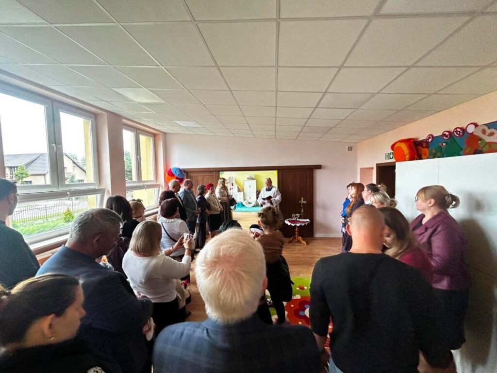 Grupa ludzi stoi w jasnej sali z jasnoróżowymi ścianami. Widoczne są balony i kolorowy dywan na podłodze. W tle ksiądz prowadzi ceremonię przy stołach.