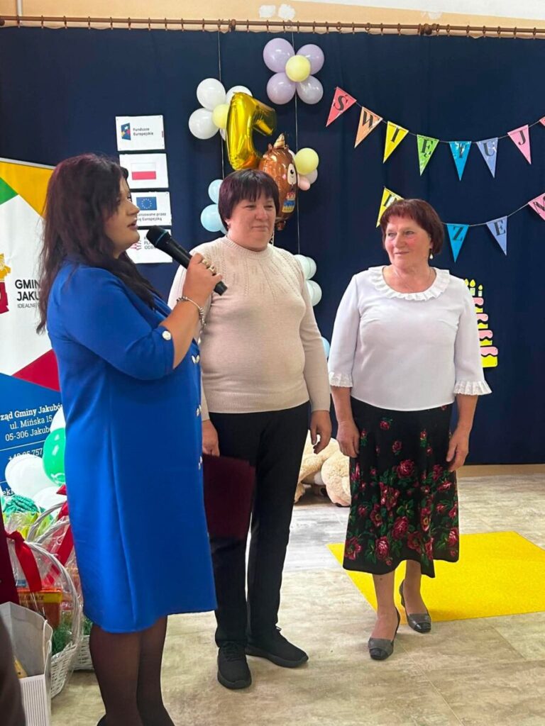 Na zdjęciu są trzy kobiety, z czego jedna trzyma mikrofon. Kobiety stoją na tle ciemnoniebieskiej zasłony z balonami i kolorową girlandą. Na podłodze jasne kafelki, a na stole widać prezenty.