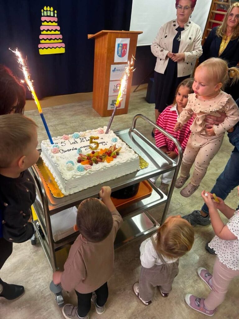 Dzieci otaczają tort z napisem 5 lat żłobka z fontannami. Tło z dekoracją przedstawiającą tort. Dorośli stoją z tyłu.