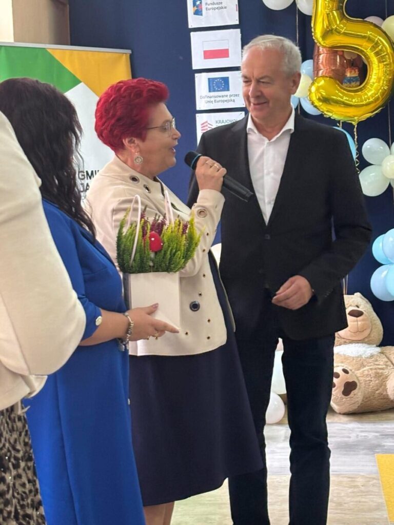 Na zdjęciu jest grupa osób, kobieta w białej marynarce mówi do mikrofonu. W tle są balony i flagi. Kolory dominujące to niebieski i żółty.