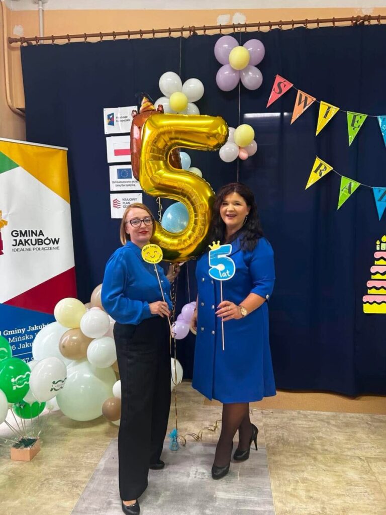 Dwie kobiety w niebieskich strojach trzymają złoty balon z cyfrą 5. Tło ozdobione balonami i kolorowymi flagami z napisami. Obok znajduje się plakat z logo Gminy Jakubów.