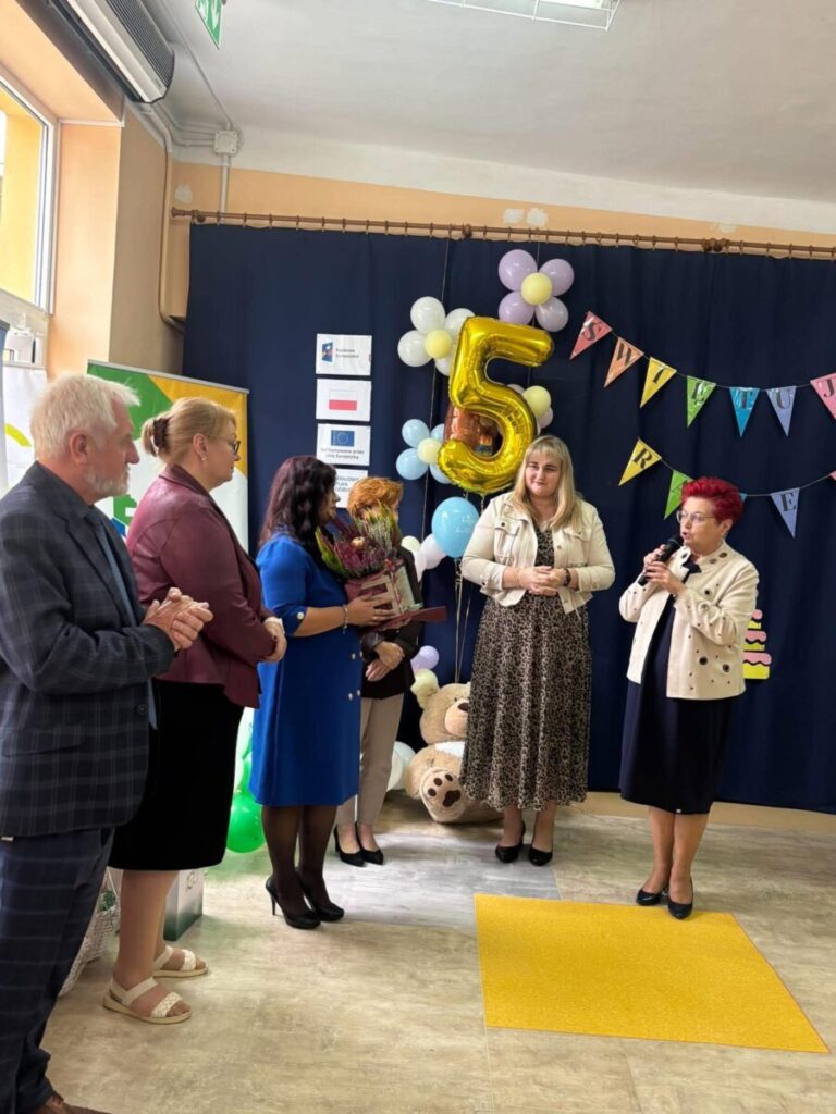 Grupa ludzi w formalnych strojach pozuje przy ozdobach na przyjęciu. W tle złoty balon z cyfrą 5 i kolorowe balony. Tło granatowe z girlandą i flagami na ścianie.