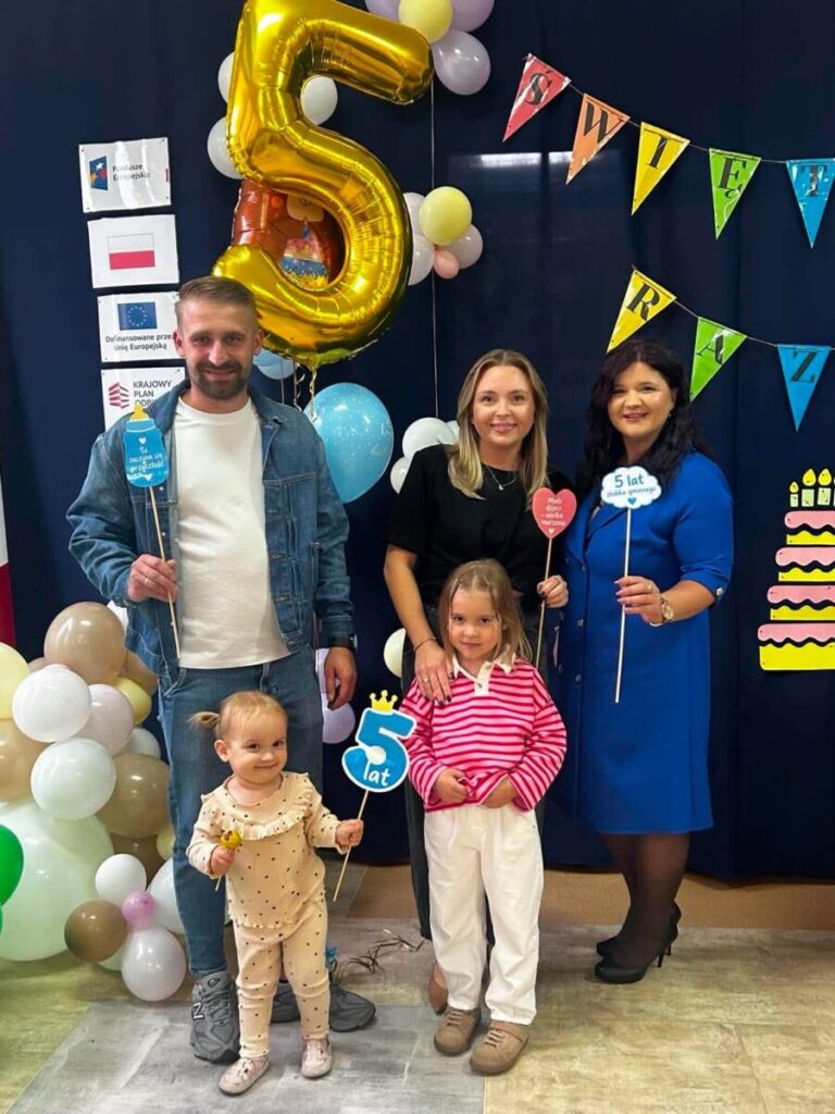 Rodzina stoi przed ciemnym tłem z kolorowymi balonami i napisem „5 lat”. Dzieci trzymają rekwizyty z liczbą 5. W tle widać dekoracje urodzinowe.