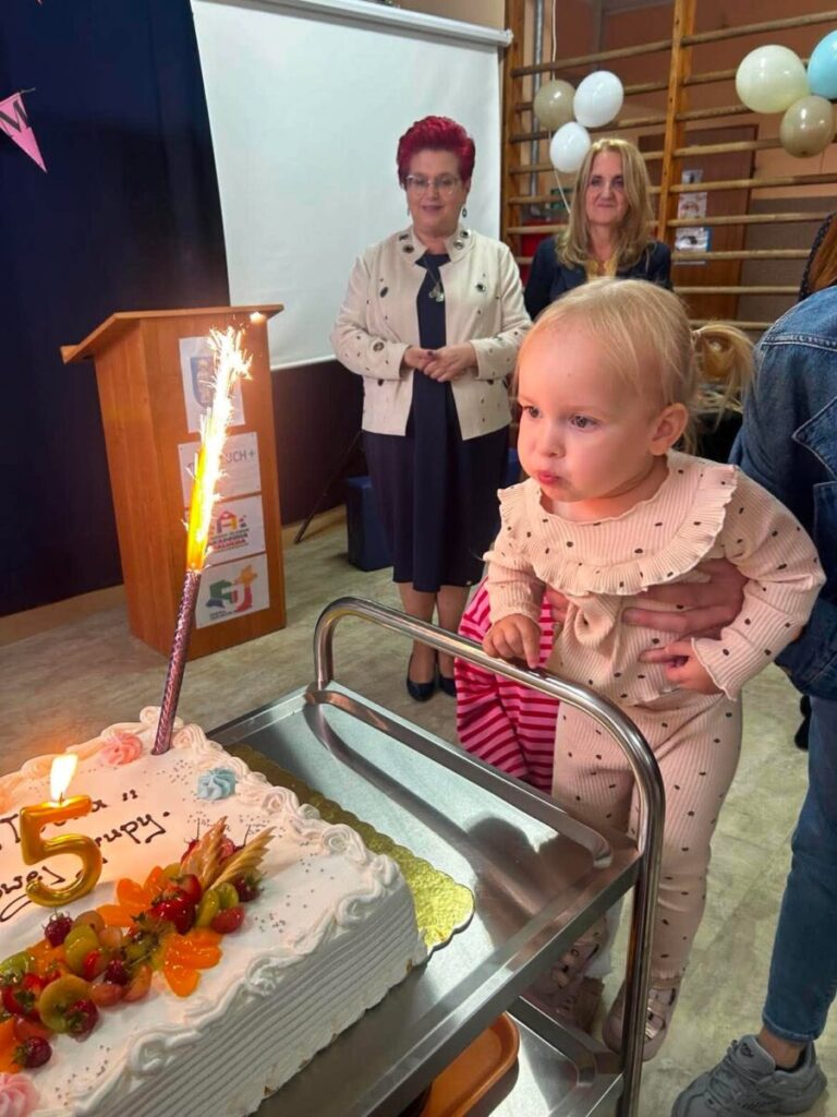 Małe dziecko w jasnym stroju stoi przy torcie z cyfrą 5 i świeczką w formie fontanny. W tle dwie dorosłe osoby, dekoracje z balonów. Pomieszczenie ma jasne ściany i drewnianą drabinkę.