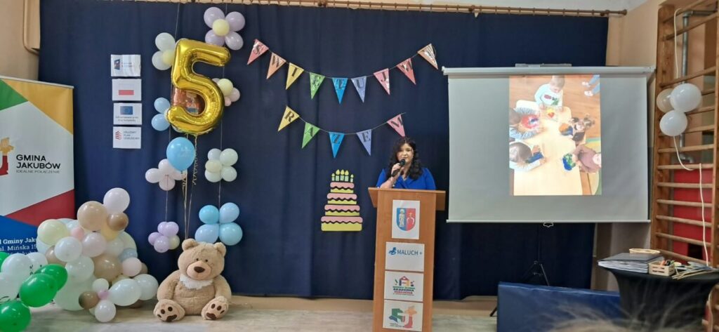 Sala udekorowana na uroczystość; duży, złoty balon w kształcie cyfry 5, kolorowe balony i plansza z projektorem.