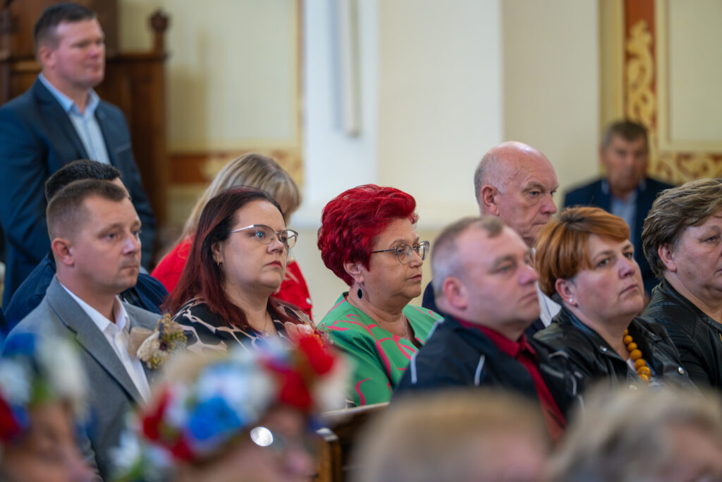 Grupa osób siedzi w sali z ozdobnymi elementami architektonicznymi. Przeważają kolory czerwony, zielony i niebieski. Na pierwszym planie kwiatowe ozdoby.