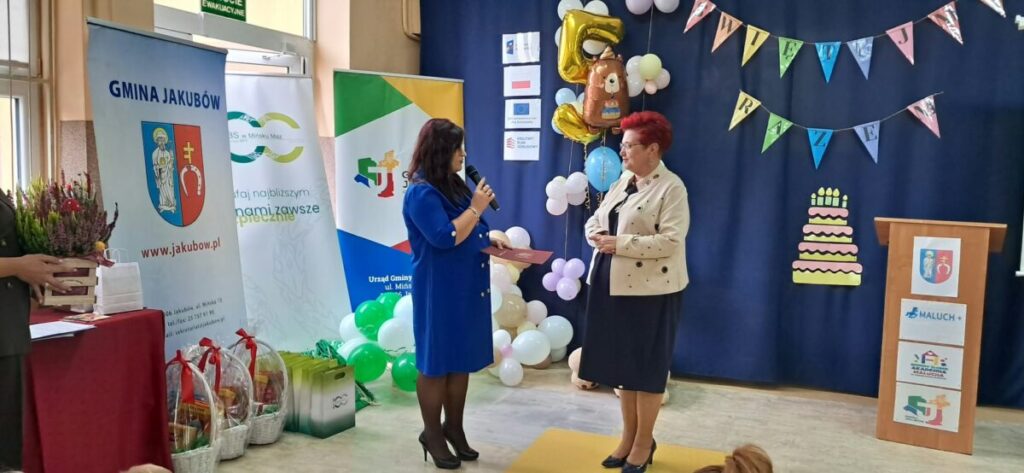 Dwóch ludzi stoi na scenie z niebieskim tłem i kolorowymi balonami. Na ścianie transparenty i diagramy, obok stojaki z logotypami. Przed nimi stół z prezentami zapakowanymi w kosze.