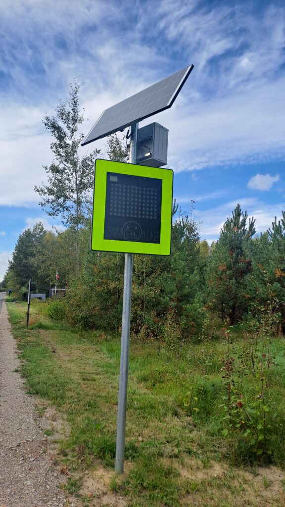 Zielona tablica zasilana panelem słonecznym stoi przy drodze. Obok znajduje się zalesiony teren i jasne, niebieskie niebo.