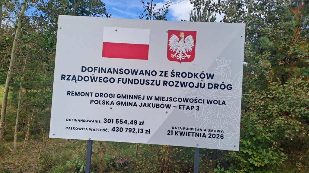 Na zdjęciu widoczna jest tablica informacyjna projektu drogowego. Ma biały kolor z flagą i godłem Polski na górze. W tle znajdują się drzewa i niebo.