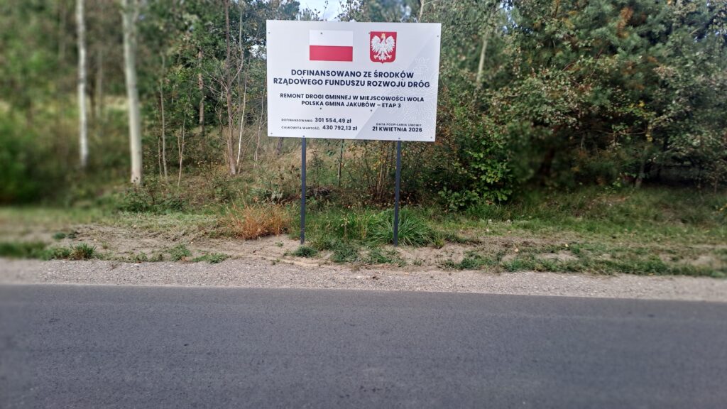 Tablica informacyjna na tle drzew informuje o dofinansowaniu remontu drogi. Widoczne barwy: biel, czerwień, zieleń. Asfaltowa nawierzchnia z przodu.
