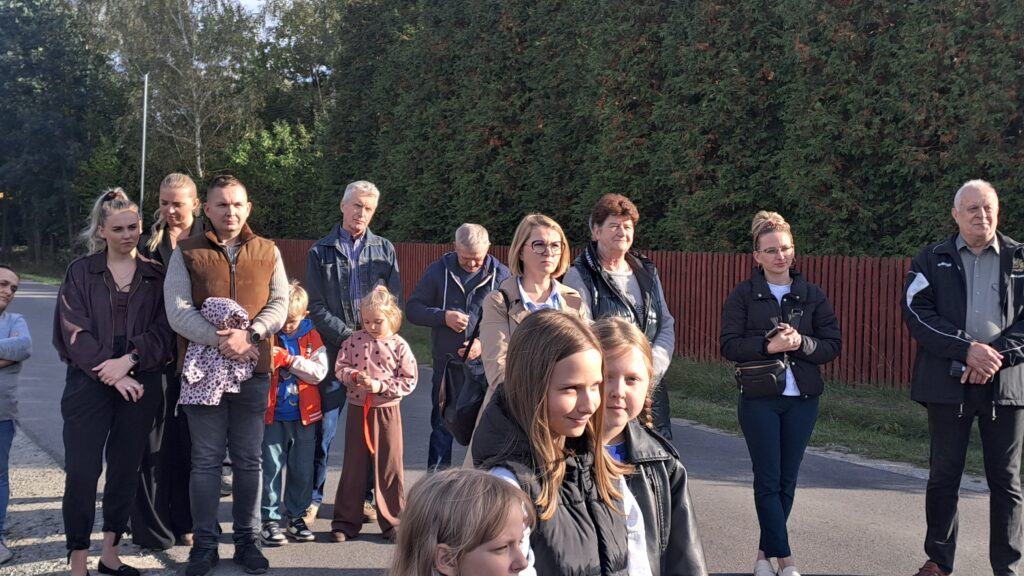 Grupa ludzi stoi na ulicy w słońcu, otoczona zielonymi drzewami i czerwonym płotem. Wśród nich są dzieci i dorośli. Każdy patrzy w stronę aparatu.