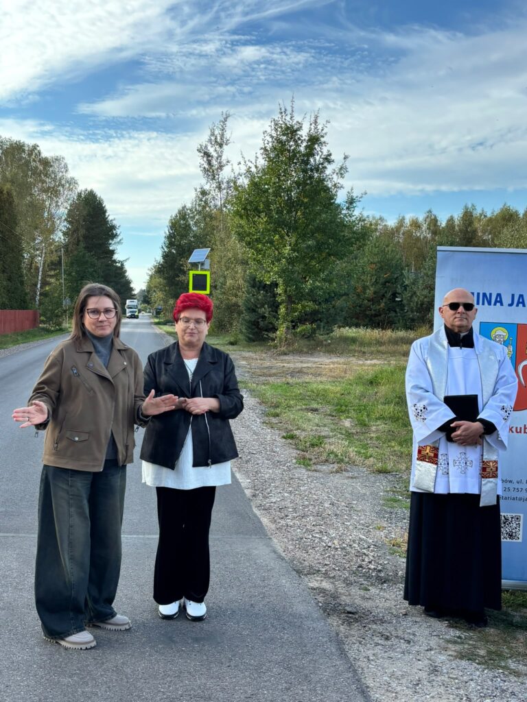 Na zdjęciu trzy osoby stoją przy drodze. Tło to zielone drzewa i błękitne niebo. Po prawej stronie widoczny jest znak z napisem.