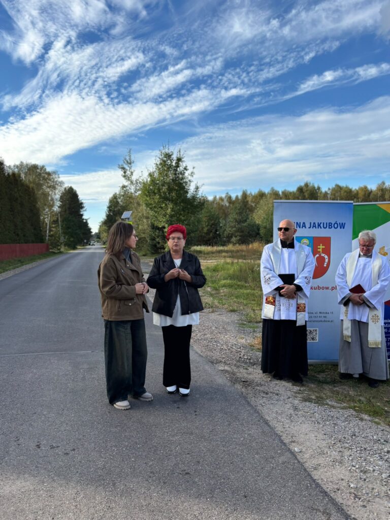 Grupa ludzi stoi przy drodze obok znaku gminy Jakubów. Są ubrani w różnorodne stroje; niebo jest błękitne z chmurami. W tle widać drzewa i zieleń.