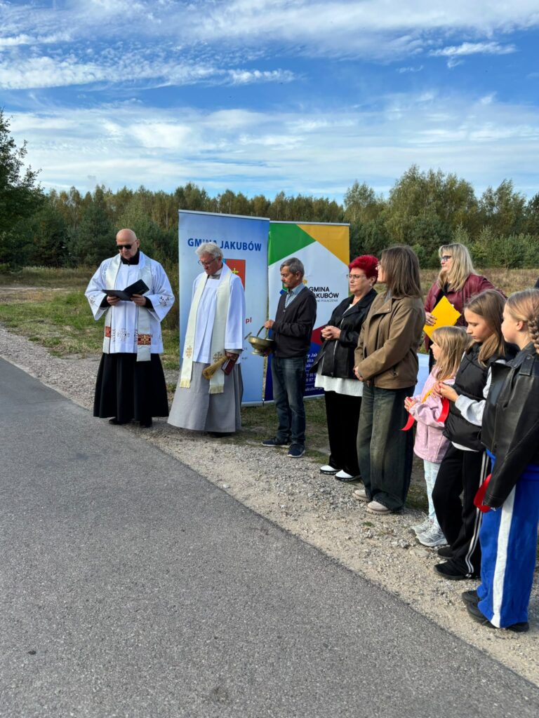 Grupa ludzi stoi na drodze przy lesie, w tym dwaj duchowni w białych i czarnych szatach. W tle widoczne są banery gminy Jakubów. Niebo jest częściowo zachmurzone.