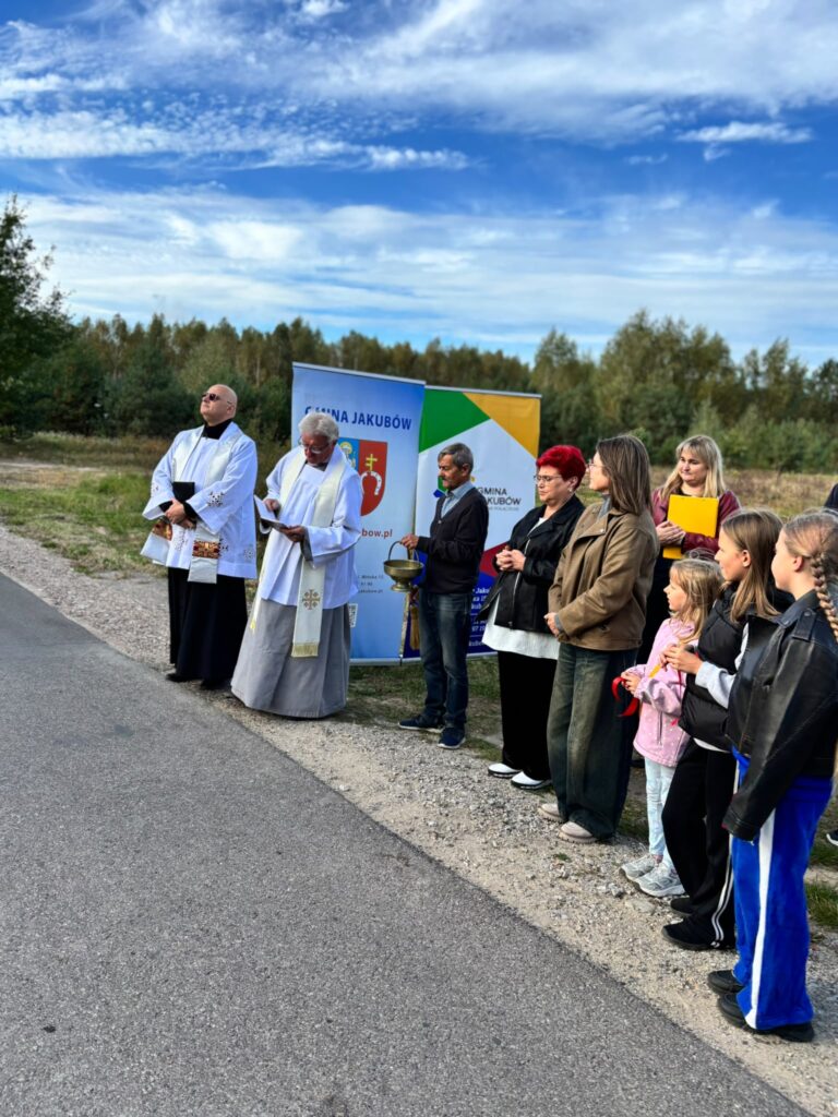 Grupa ludzi stoi na poboczu drogi obok banerów. Dwie osoby są ubrane w białe szaty, tło stanowi zalesiony krajobraz pod niebieskim niebem. Widać czerwone wstążki i złotą misę trzymaną przez jedną z osób